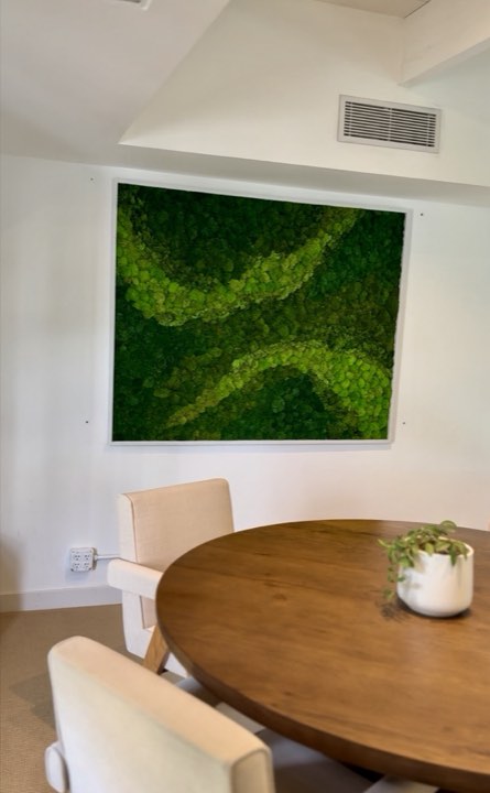Large framed moss art with a flowing light-green swirl pattern above a round wood dining table